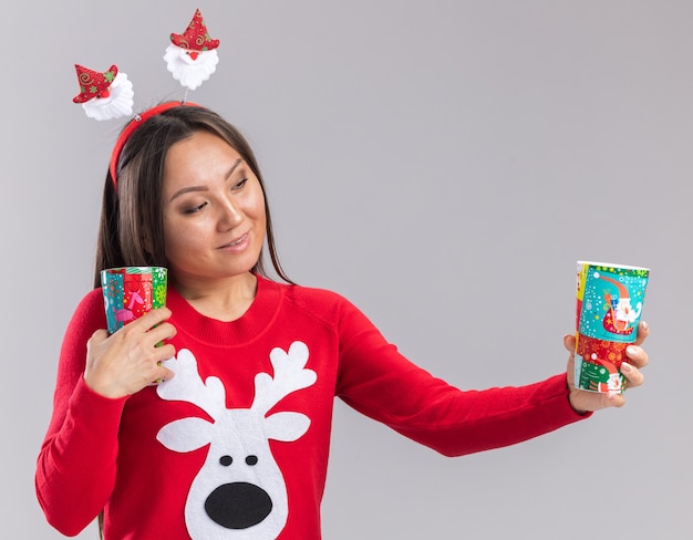 Erfreutes junges asiatisches Mädchen, das Weihnachtshaarbügel mit Pullover hält und Weihnachtsbecher lokalisiert auf weißem Hintergrund hält