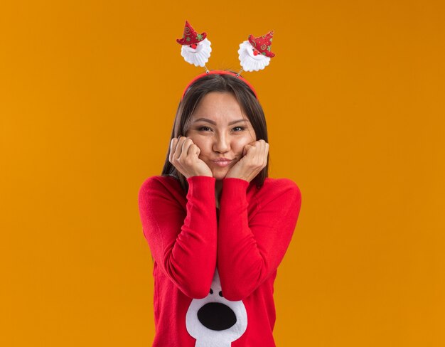 Erfreutes junges asiatisches Mädchen, das Weihnachtshaarreifen mit Pullover trägt, der Fäuste auf Wangen setzt, die auf orange Wand lokalisiert werden