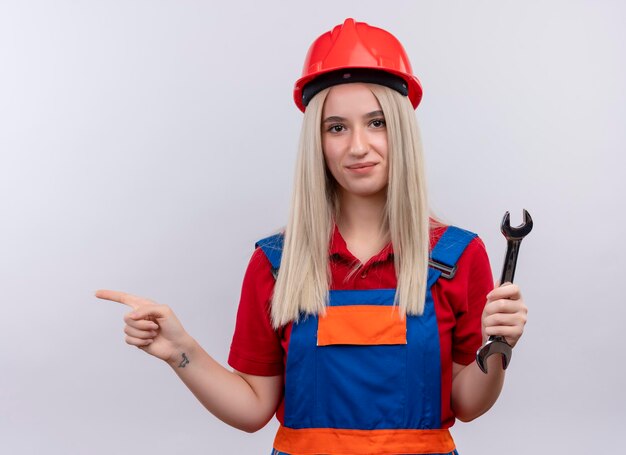 Erfreutes junges blondes Ingenieur-Baumeistermädchen in der Uniform, die den offenen Schraubenschlüssel hält, der auf der linken Seite auf isolierten weißen Raum zeigt