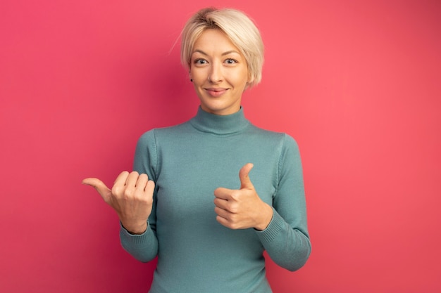 Erfreutes junges blondes Mädchen, das auf die Seite zeigt, die Daumen nach oben zeigt