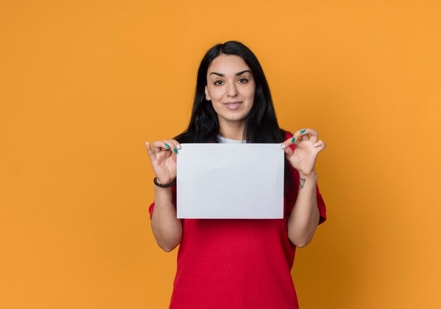 Erfreutes junges brünettes kaukasisches Mädchen, das rotes Hemd trägt, hält Papierblatt, das auf orange Wand lokalisiert schaut