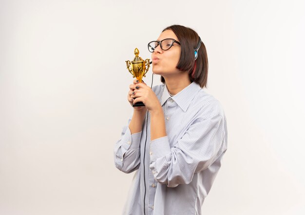Erfreutes junges Callcenter-Mädchen, das Brille und Headset küsst Gewinnerpokal mit geschlossenen Augen lokalisiert auf weißem Hintergrund mit Kopienraum