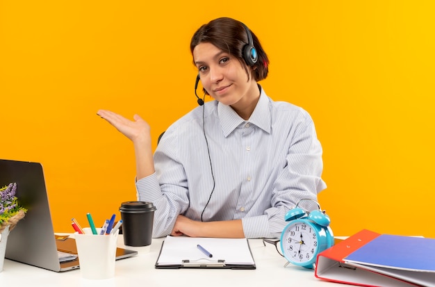 Erfreutes junges Callcenter-Mädchen, das Headset trägt, das am Schreibtisch sitzt und leere Hände lokalisiert auf orange Hintergrund zeigt