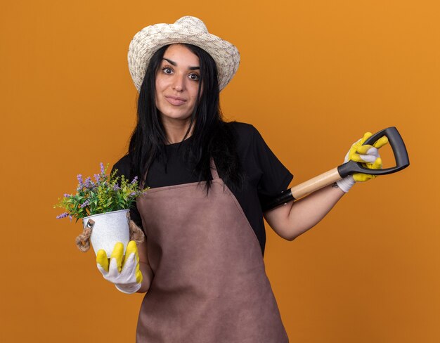 Erfreutes junges Gärtnermädchen in Uniform und Hut mit Gärtnerhandschuhen, das Spaten hinter dem Rücken hält und Blumentopf mit Blick auf die Vorderseite isoliert auf oranger Wand