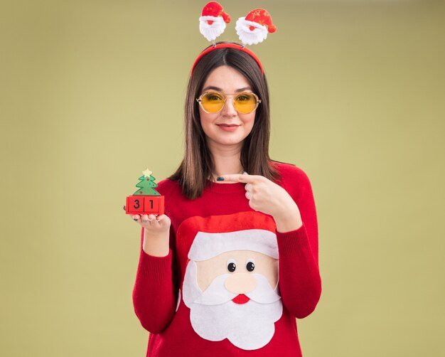 Erfreutes junges hübsches kaukasisches Mädchen, das Weihnachtsmann-Pullover und Stirnband mit Brille hält, die auf Weihnachtsbaumspielzeug mit Datum hält und zeigt