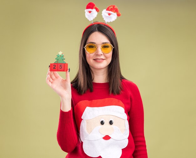 Erfreutes junges hübsches kaukasisches Mädchen, das Weihnachtsmann-Pullover und Stirnband mit Gläsern hält, die Weihnachtsbaumspielzeug mit Datum betrachten Kamera betrachten, die auf olivgrünem Hintergrund lokalisiert wird