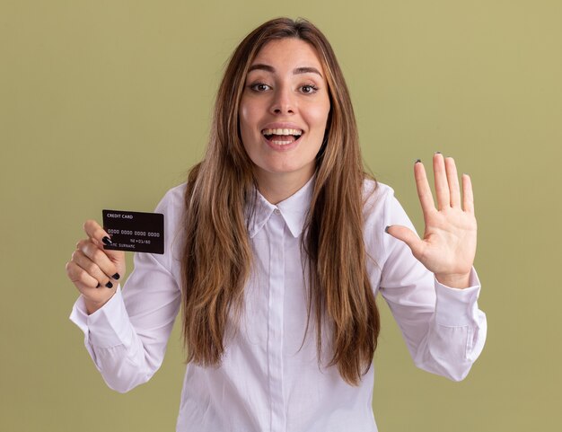 Erfreutes junges hübsches kaukasisches Mädchen hält Kreditkarte und gestikuliert fünf auf Olivgrün