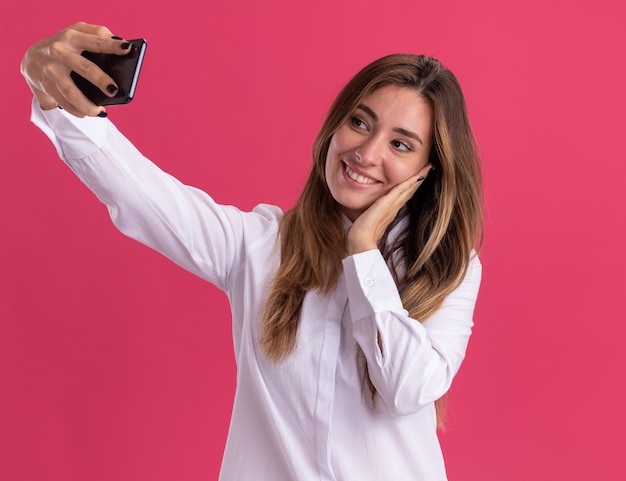 Erfreutes junges hübsches kaukasisches Mädchen legt die Hand auf das Gesicht, das das Telefon hält und betrachtet, das Selfie macht