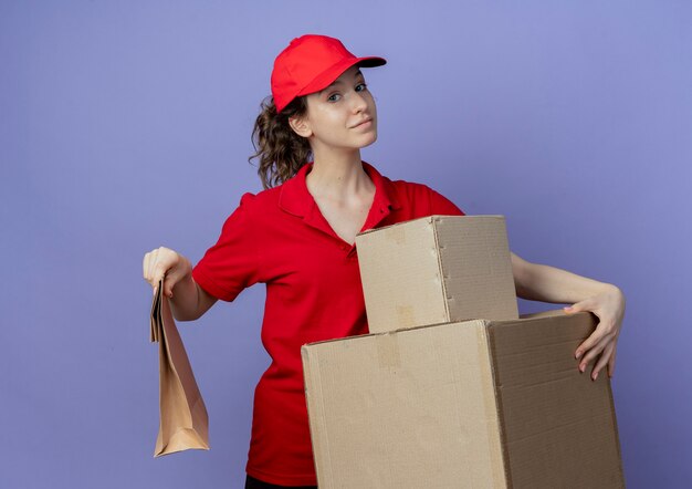 Erfreutes junges hübsches Liefermädchen, das rote Uniform und Kappe hält, die Kartonkästen und Papierpaket lokalisiert auf lila Hintergrund hält