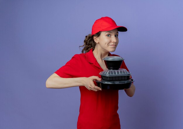 Erfreutes junges hübsches Liefermädchen, das rote Uniform und Kappe hält, die Lebensmittelbehälter lokalisiert auf lila Hintergrund mit Kopienraum hält