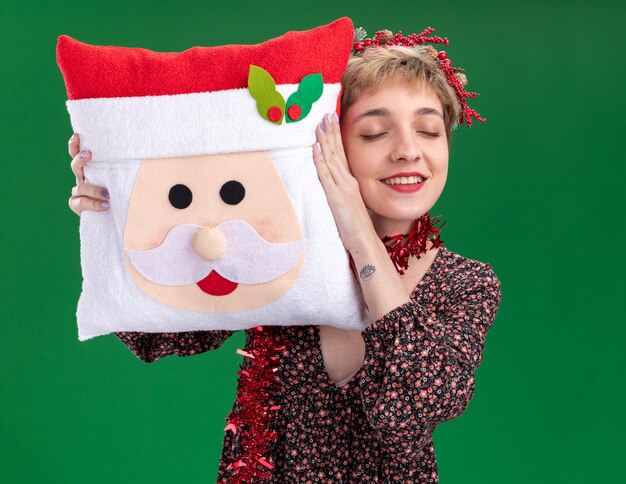 Erfreutes junges hübsches Mädchen, das Weihnachtskopfkranz und Lametta-Girlande um den Hals trägt, hält Weihnachtsmann-Kissen, das Kopf berührt, der mit geschlossenen Augen auf grünem Hintergrund lokalisiert lächelt