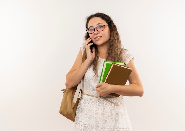 Erfreutes junges hübsches Schulmädchen, das Brille und Rückentasche hält Bücher hält, die am Telefon sprechen, lokalisiert auf weißer Wand