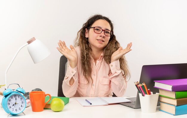 Erfreutes junges hübsches Schulmädchen, das eine Brille trägt, die mit Schulwerkzeugen am Schreibtisch sitzt und ihre Hausaufgaben macht, die leere Hände zeigen, die auf weißer Wand isoliert werden