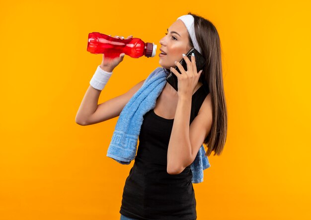 Erfreutes junges hübsches sportliches Mädchen, das Stirnband und Armband trägt und versucht, Wasser aus der Flasche zu trinken und am Telefon zu sprechen, das Seite betrachtet, die auf orange Raum lokalisiert wird