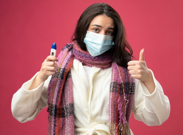 Kostenloses Foto erfreutes junges kaukasisches krankes mädchen, das robe und schal mit maske hält, die thermometer hält daumen oben auf purpurroter wand lokalisiert