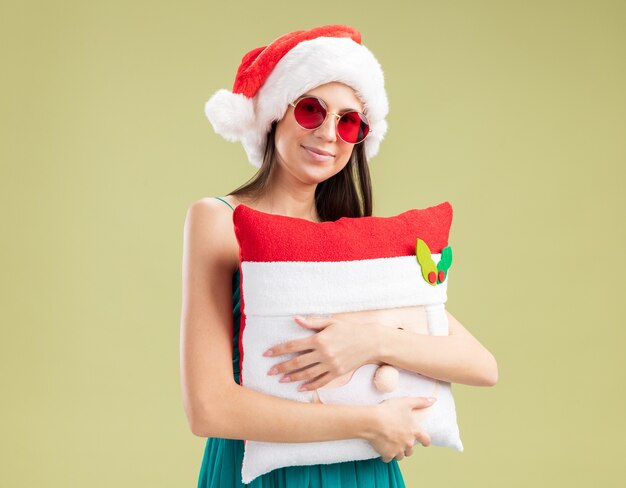 Erfreutes junges kaukasisches Mädchen in Sonnenbrille mit Weihnachtsmütze umarmt Weihnachtsmannkissen