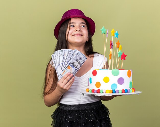 erfreutes junges kaukasisches Mädchen mit lila Partyhut, das Geburtstagskuchen und Geld hält, das auf der Seite isoliert auf olivgrüner Wand mit Kopierraum schaut
