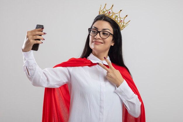 Erfreutes junges kaukasisches Superheldenmädchen, das Brille und Krone tut Friedenszeichen nimmt, das selfie lokalisiert auf weißem Hintergrund mit Kopienraum nimmt