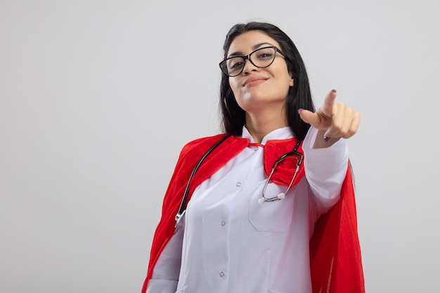 Erfreutes junges kaukasisches Superheldenmädchen, das Brille und Stethoskop trägt und auf Kamera zeigt, lokalisiert auf weißem Hintergrund mit Kopienraum