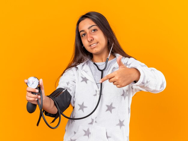 Erfreutes junges krankes Mädchen, das ihren eigenen Druck mit dem auf Gelb isolierten Blutdruckmessgerät misst