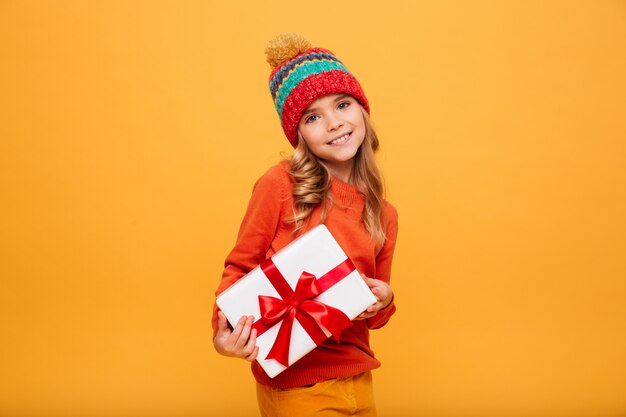 Erfreutes junges Mädchen in der Strickjacke und in Hut, die Geschenkbox halten und die Kamera über Orange betrachten