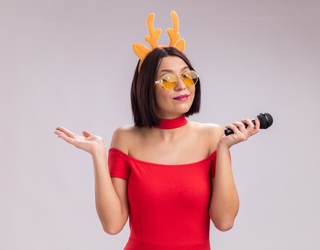 Erfreutes junges Mädchen mit Rentiergeweih-Stirnband und Brille mit Mikrofon und Blick in die Kamera mit leerer Hand isoliert auf weißem Hintergrund