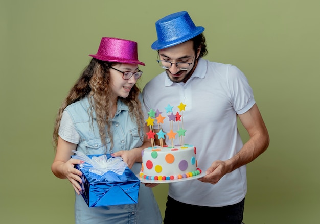 Erfreutes junges Paar, das rosa und blauen Hutmädchen hält Geschenkbox und Kerl hält und mit Mädchen an Geburtstagstorte schaut