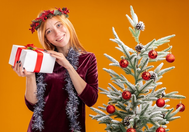 erfreutes junges schönes Mädchen, das in der Nähe einen Weihnachtsbaum trägt und ein rotes Kleid und einen Kranz mit Girlande am Hals trägt, die eine Geschenkbox isoliert auf einer orangefarbenen Wand hält