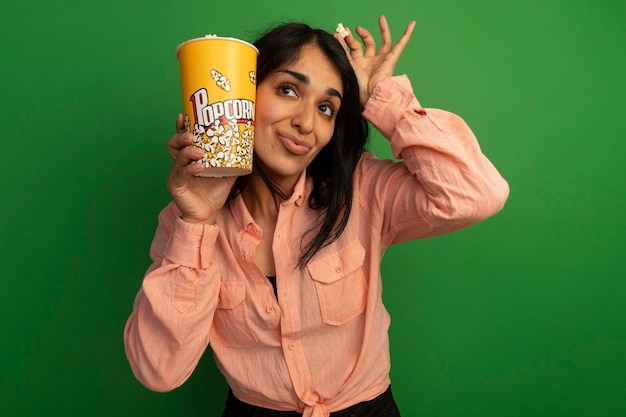 Erfreutes junges schönes Mädchen, das rosa T-Shirt hält Eimer Popcorn mit Popcorn Frieden um Gesicht lokalisiert auf grüner Wand hält