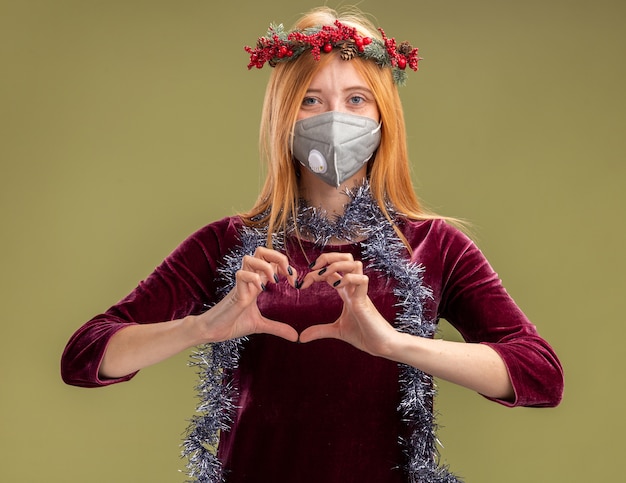 Kostenloses Foto erfreutes junges schönes mädchen, das rotes kleid mit kranz und medizinischer maske mit girlande am hals trägt, die herzgeste lokalisiert auf olivgrünem hintergrund zeigt