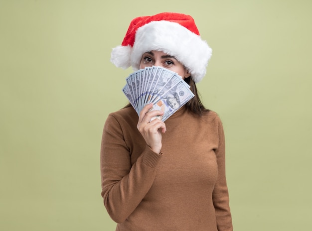 Erfreutes junges schönes Mädchen, das Weihnachtshut bedecktes Gesicht mit Bargeld lokalisiert auf olivgrünem Hintergrund trägt