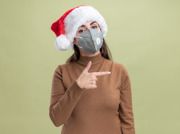 Erfreutes junges schönes Mädchen, das Weihnachtshut mit medizinischen Maskenpunkten an der Seite lokalisiert auf olivgrünem Hintergrund mit Kopienraum trägt