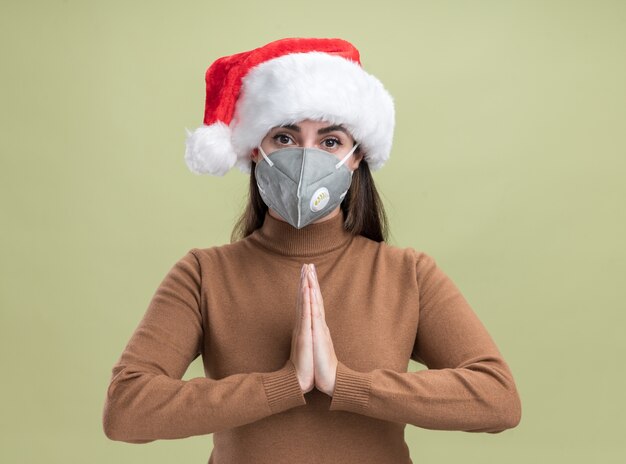 Erfreutes junges schönes Mädchen, das Weihnachtshut mit medizinischer Maske trägt, die Gebetsgeste auf olivgrünem Hintergrund zeigt