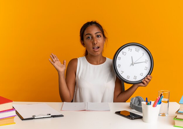 Erfreutes junges Schulmädchen, das am Schreibtisch mit Schulwerkzeugen sitzt, die Wanduhr halten und Hand auf Orange ausbreiten