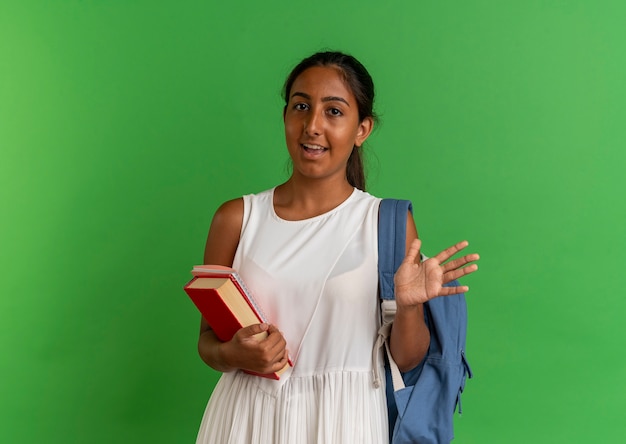 Erfreutes junges Schulmädchen, das Rückentasche hält Buch mit Notizbuch und Hand auf Grün ausbreitet