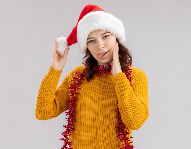 Erfreutes junges slawisches Mädchen mit Weihnachtsmütze und mit Girlande um den Hals streckt die Zunge heraus und legt die Hand auf das Gesicht isoliert auf weißer Wand mit Kopierraum