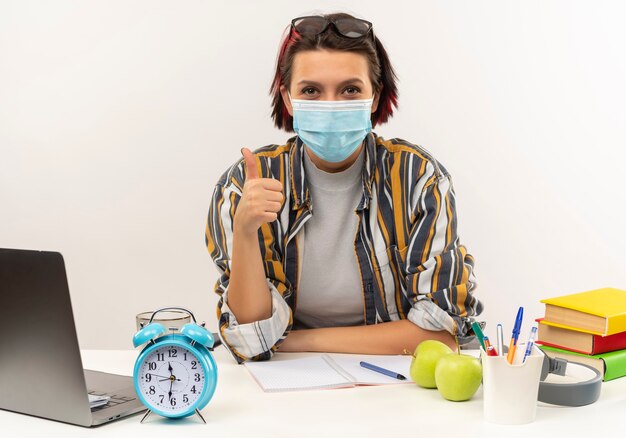 Erfreutes junges Studentenmädchen, das Brille auf Kopf und Maske trägt, die am Schreibtisch sitzt, zeigt Daumen oben isoliert auf weißer Wand