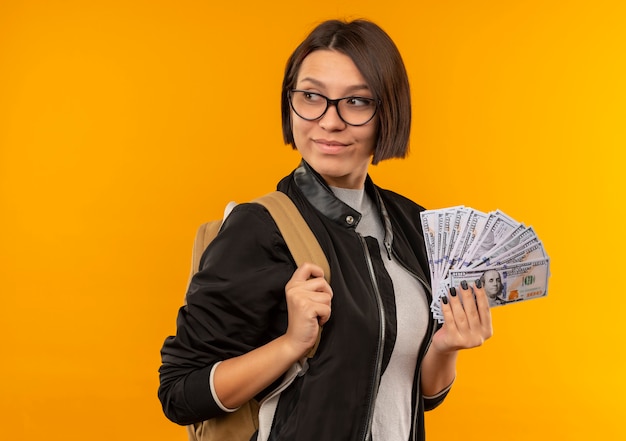 Erfreutes junges Studentenmädchen, das Brille und Rückentasche hält, die Geld hält, das Seite auf orange Wand lokalisiert betrachtet