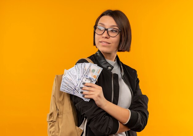 Erfreutes junges Studentenmädchen, das Brille und Rückentasche trägt, die mit geschlossener Haltung hält, die Geld hält, das Seite lokalisiert auf orange Wand betrachtet