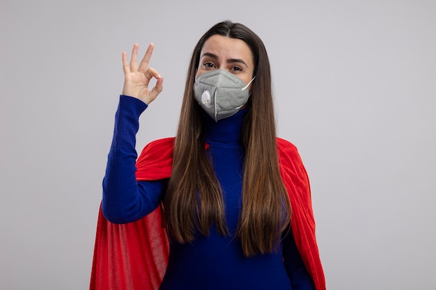 Kostenloses Foto erfreutes junges superheldenmädchen, das medizinische maske trägt, die okay geste lokalisiert auf weißem hintergrund zeigt