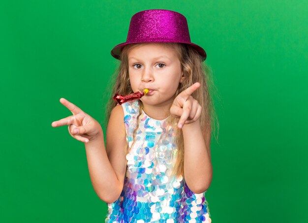 erfreutes kleines blondes Mädchen mit lila Partyhut bläst Partypfeife und gestikuliert Siegeszeichen isoliert auf grüner Wand mit Kopierraum