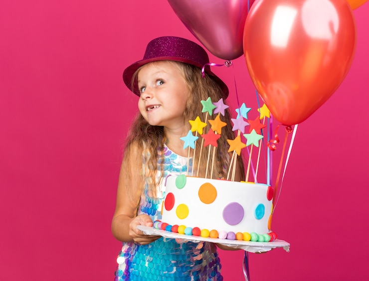 Kostenlose Foto erfreutes kleines blondes mädchen mit lila partyhut, der heliumballons und geburtstagstorte hält, die auf rosa wand mit kopienraum lokalisiert werden