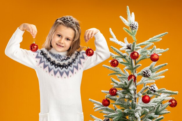 Erfreutes kleines Mädchen, das nahe Weihnachtsbaum steht, der Tiara mit Girlande am Hals hält, der Weihnachtskugeln lokalisiert auf orange Hintergrund hält