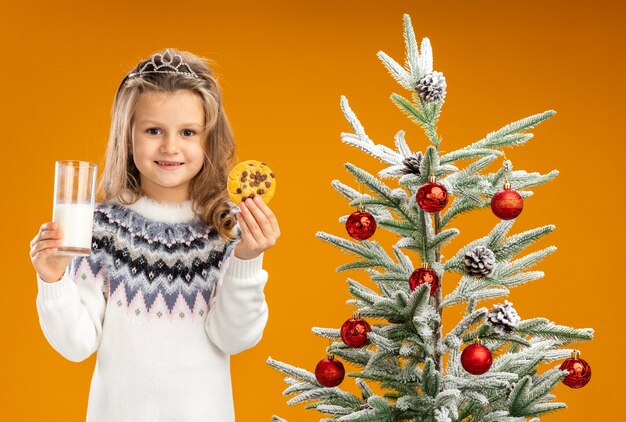 Erfreutes kleines Mädchen, das nahe Weihnachtsbaum steht, der Tiara mit Girlande am Hals hält Glas der Milch mit Keksen lokalisiert auf orange Hintergrund trägt