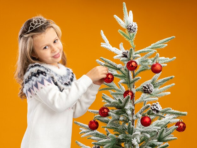 Erfreutes kleines Mädchen, das nahe Weihnachtsbaum steht, der Tiara mit Girlande am Hals trägt, hängt Spielzeug auf dem Baum, der auf orange Hintergrund lokalisiert wird