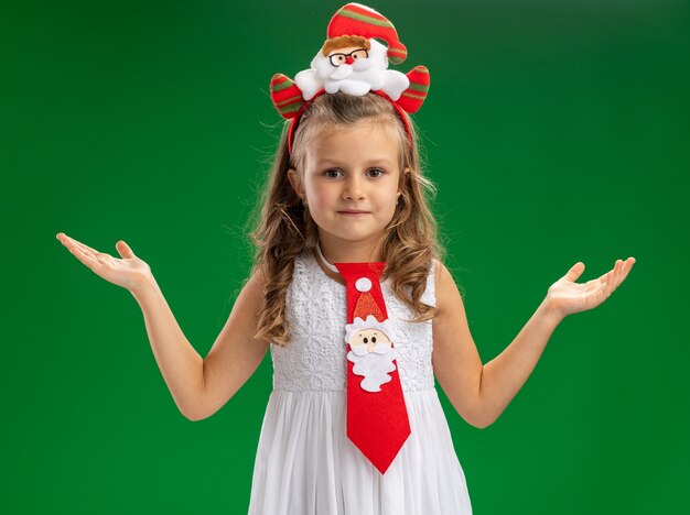 Erfreutes kleines Mädchen, das Weihnachtshaarreifen mit Krawattenverbreitungshänden lokalisiert auf grünem Hintergrund trägt
