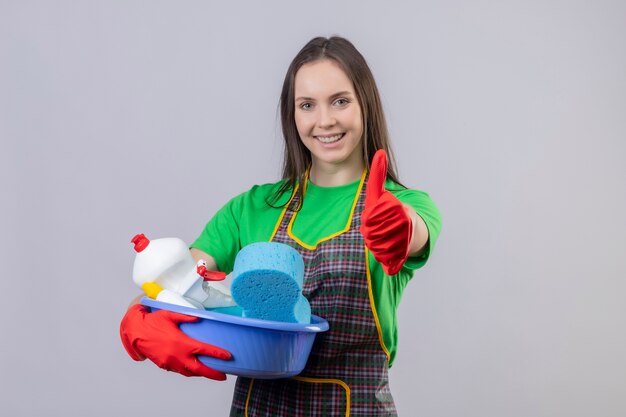 Erfreutes Reinigen des jungen Mädchens, das Uniform in den roten Handschuhen hält, die Reinigungswerkzeuge ihren Daumen oben auf lokalisiertem weißem Hintergrund halten