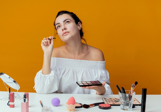 Erfreutes schönes Mädchen sitzt am Tisch mit Make-up-Werkzeugen, die Lidschattenpalette und Make-up-Pinsel lokalisiert auf orange Wand halten