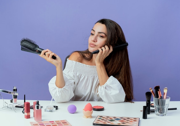 Erfreutes schönes Mädchen sitzt am Tisch mit Make-up-Werkzeugen hält Haarkämme, die Seite lokalisiert auf lila Wand betrachten