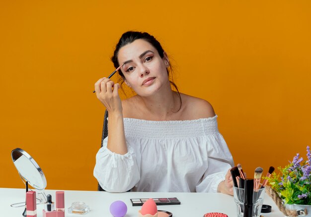 Erfreutes schönes Mädchen sitzt am Tisch mit Make-up-Werkzeugen hält Make-up-Pinsel, der isoliert auf orange Wand schaut